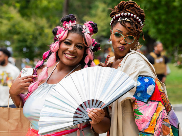 Close-up of two beautiful Black women posing wearing colorful clothing. One holds a large white folding fan.
